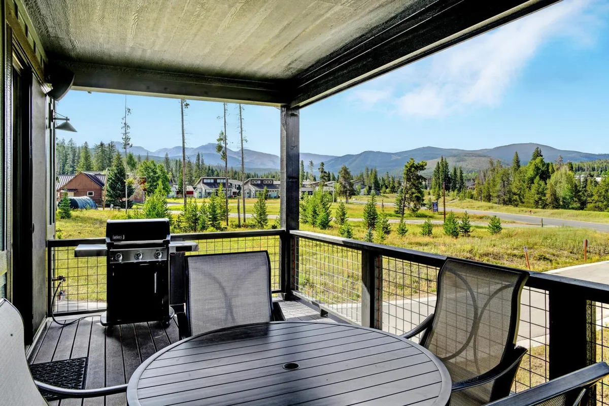 The image features the upper deck with a dining table, deck railing, and a stunning mountain view. The setting is perfect for outdoor dining, morning coffees, or watching the sky change color at sunset, offering seamless continuity from the great room to the outdoors.