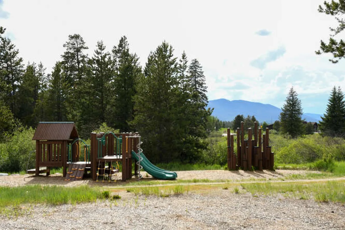 A lively playground surrounded by trees, complete with modern slides and climbing structures. Children are seen playing and laughing, while families gather nearby for picnics and outdoor games under the sunshine.