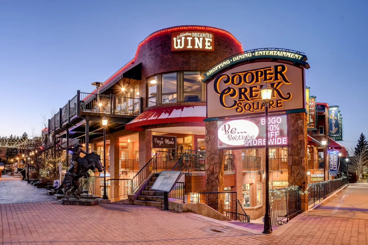 A bustling outdoor scene in downtown Winter Park’s Cooper Creek Square, featuring a prominent sign, outdoor seating areas, and local shops. People are seen dining, shopping, and socializing, with a lively yet relaxed mountain town vibe.