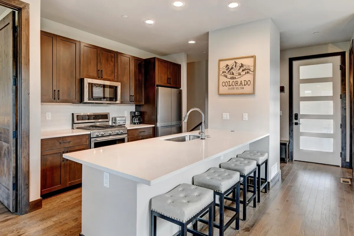 A warmly lit open-concept kitchen with a substantial island bar and stools, wooden custom cabinetry, modern appliances, and art on the walls. The space is bustling, ideal for cooking, entertaining, and gathering for meals or casual family activities.