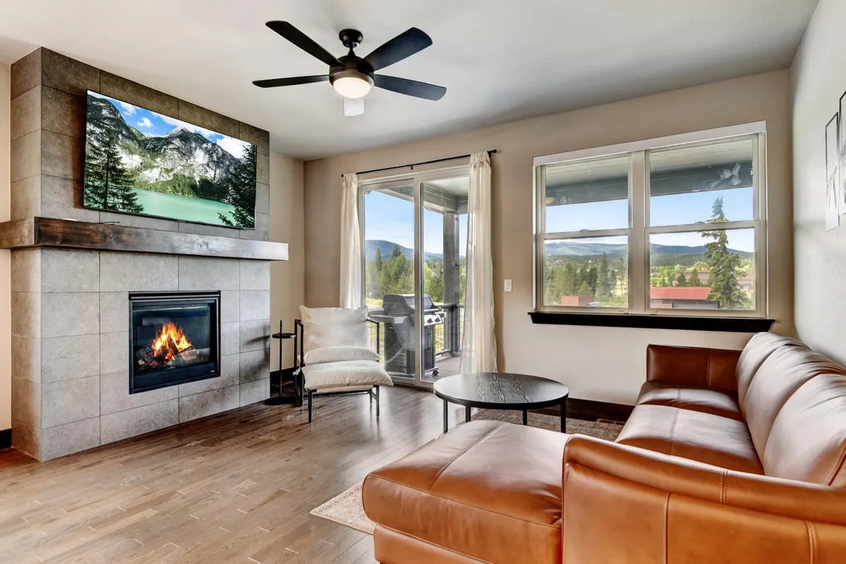 This image showcases the heart of the great room, featuring a leather sofa facing a large fireplace with a TV mounted above, a wall of windows framing expansive mountain views, and an outdoor balcony just beyond. Plush furnishings and a cozy ambiance radiate warmth, perfect for gathering and relaxing with loved ones.