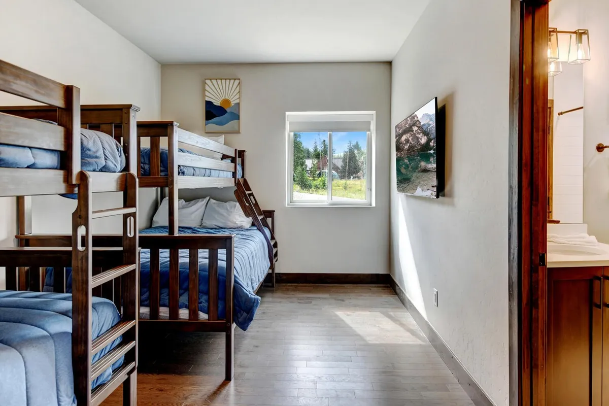 A cozy bunk room featuring bunk beds with soft bedding, a window for natural light, playful artwork, and a television. The room radiates warmth and provides a welcoming environment for sleep, play, and relaxation.
