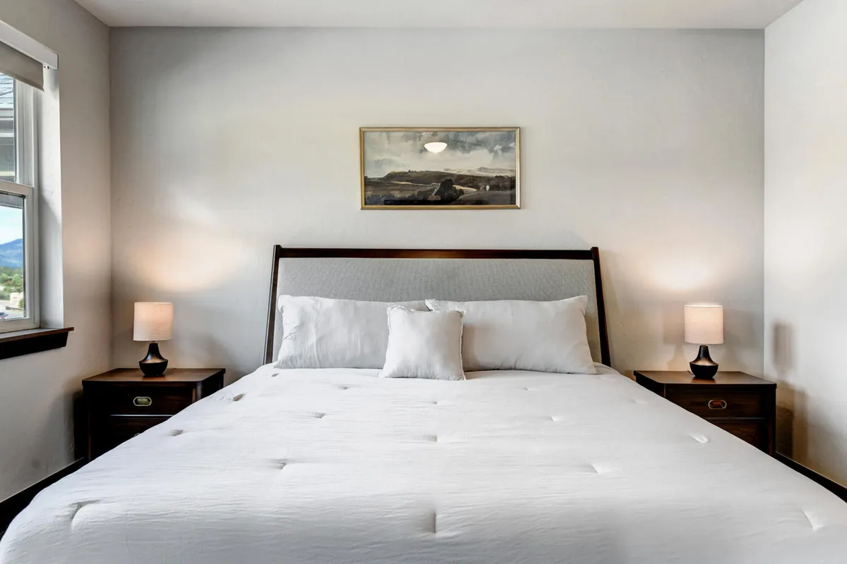 A serene master bedroom featuring a large bed with soft-toned bedding, flanked by nightstands and a calming piece of art above the headboard. Light pours in through a window, imbuing the room with a sense of peacefulness and privacy. Subtle touches of luxury are evident in the quality bedding and thoughtfully curated decor.