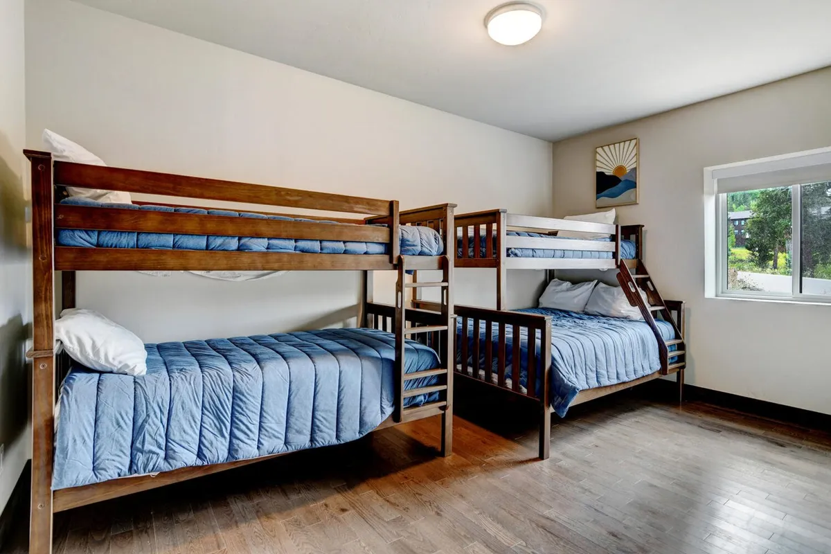 A playful and inviting bunk room with sturdy wooden twin-over-twin and twin-over-full bunk beds, dressed in soft bedding. Natural light streams through a window, brightening the space and creating a fun, camp-like atmosphere perfect for kids and friends.