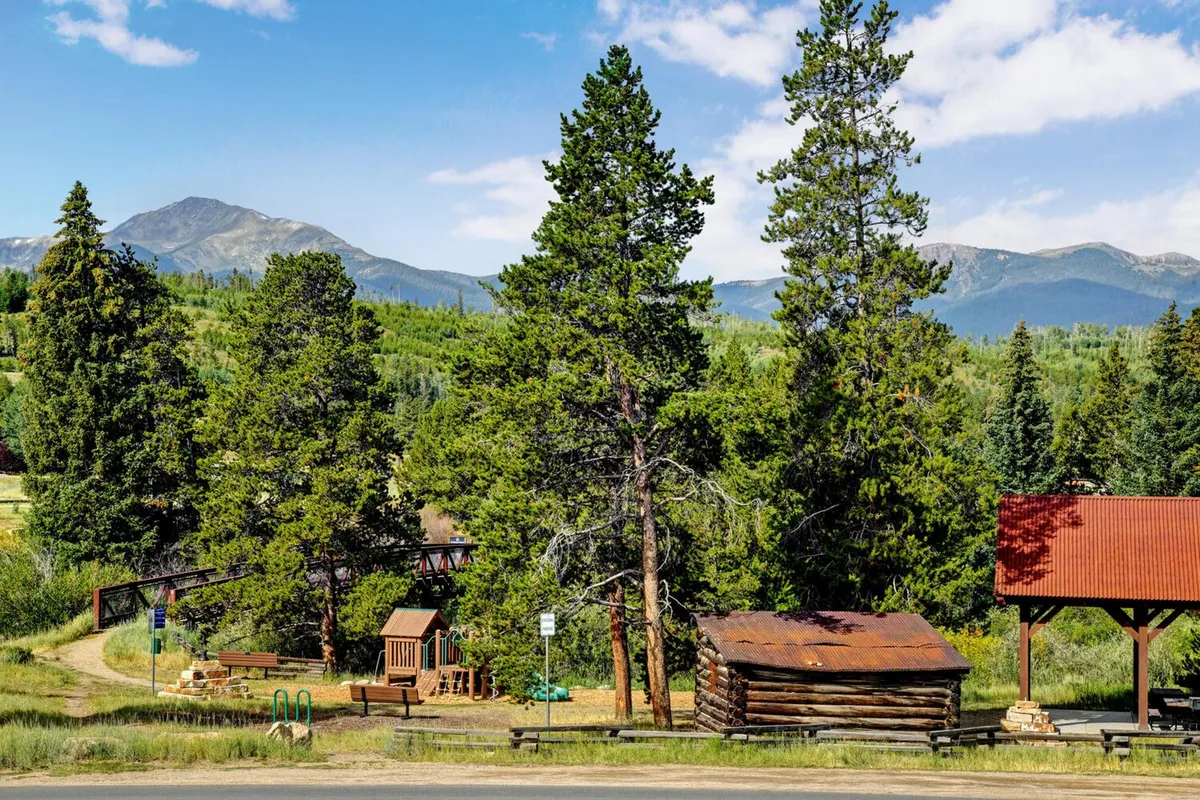 A rustic cabin sits among tall evergreens, with a family-friendly playground area and mountains rising in the background. The space is lively and inviting, suggesting opportunities for outdoor play, picnicking, and nature walks for guests of all ages.
