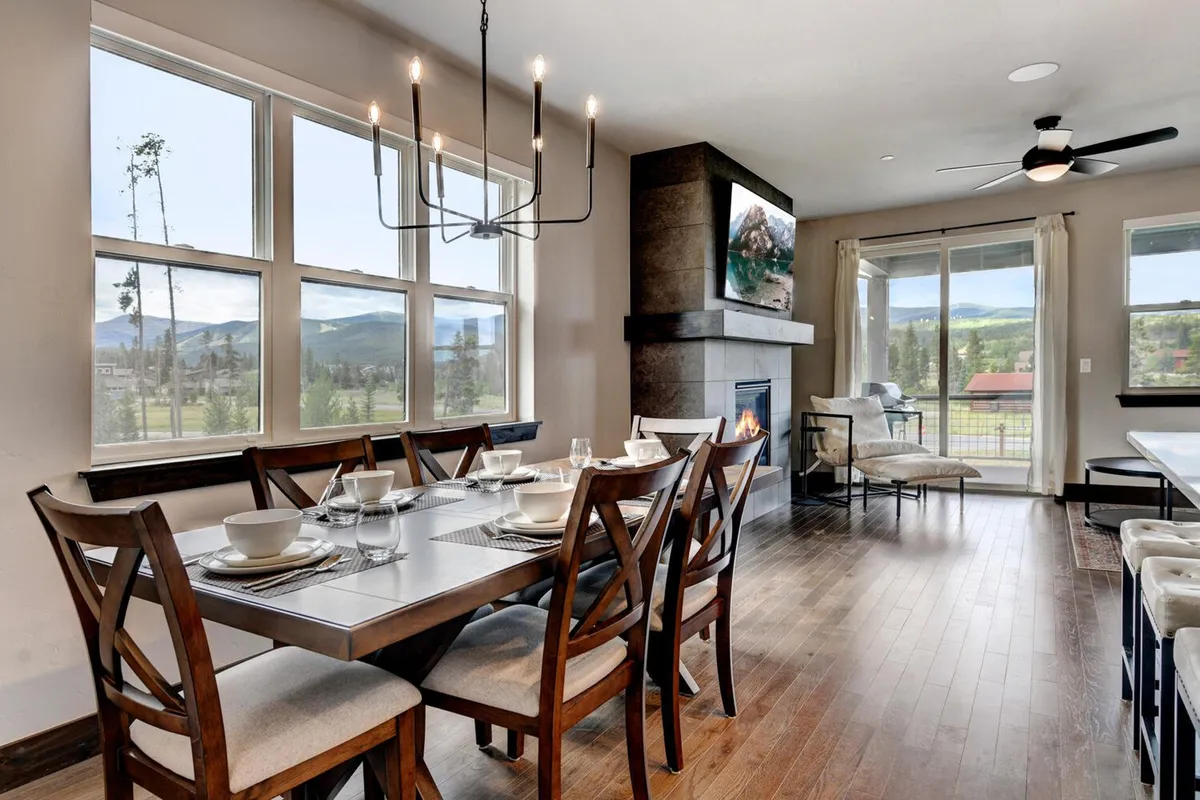 A beautifully set custom wood dining table is positioned near large windows, with a fireplace and chandelier adding warmth and elegance. The setting exudes an inviting, upscale mountain décor—ideal for shared meals, family dinners, and special celebrations, all while soaking in sunset views.