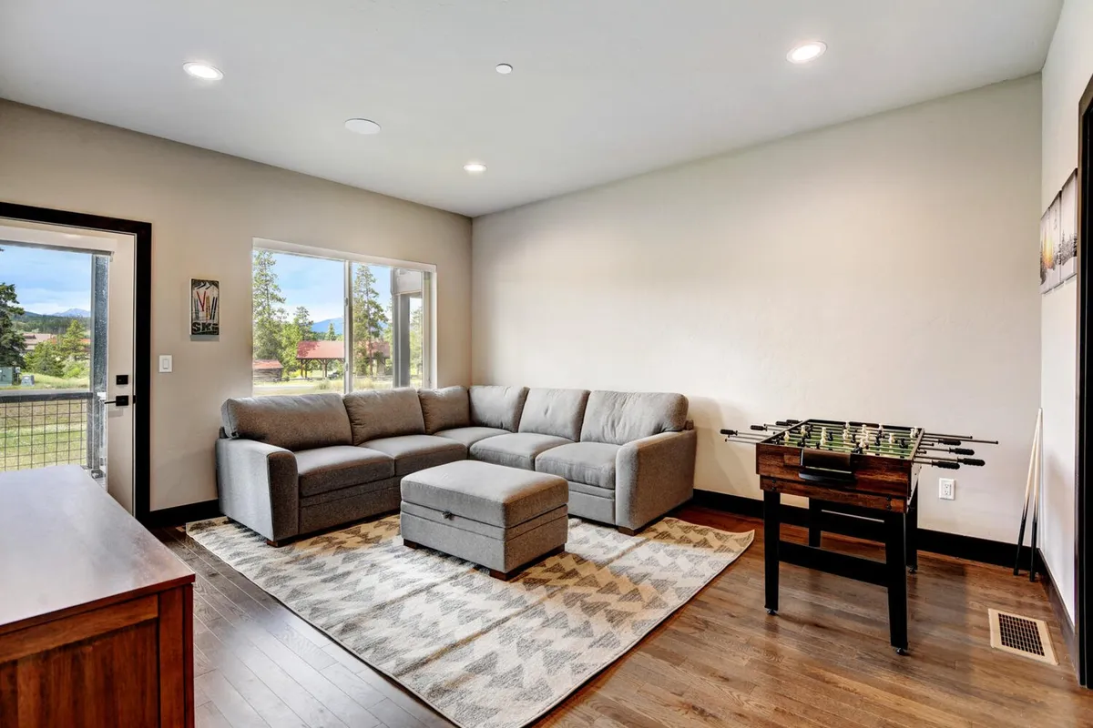A bright, inviting game room featuring a sectional sofa, a foosball table, and a large window letting in plenty of natural light. The space is arranged for both competitive fun and relaxed socializing, with enough seating for families or groups of friends to gather comfortably.